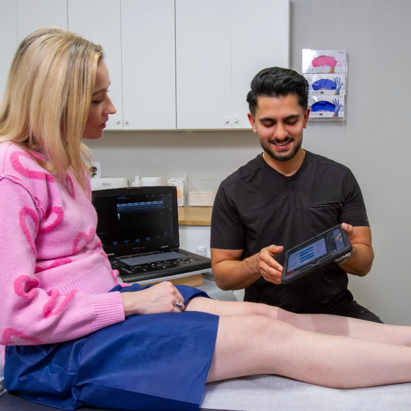 VTC_DrMehta_Lifestyle_1_optimized Dr. Akash Mehta conducts a Duplex Ultrasound examination of the patient's lower leg and foot to assess for Venous Reflux and Chronic Venous Insufficiency. The provider uses a portable Ultrasound Machine while an Ultrasound Machine with imaging software is visible on the desk in the background of this modern clinical examination room.