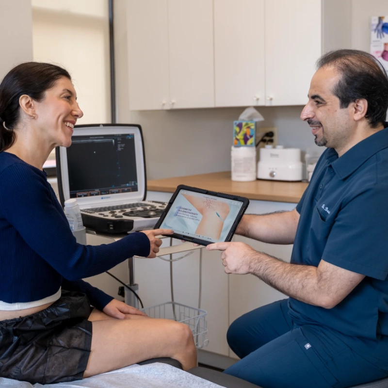 DrBasiri_Lifestyle_2__optimized Dr. Mohsen Basiri consults with a patient in a clinical setting, reviewing educational material on a tablet device regarding venous treatment options. The modern office features an Ultrasound Machine visible in the background, demonstrating a patient consultation about vein health and potential treatment plans such as Duplex Ultrasound, Sclerotherapy, or Radiofrequency Ablation.